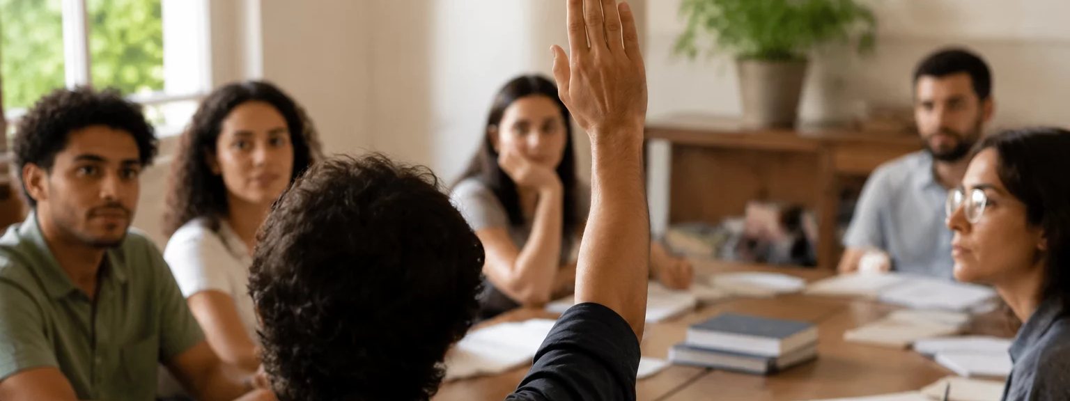 Participante com a mão erguida pedindo a vez, à mesa coberta de folhas escritas